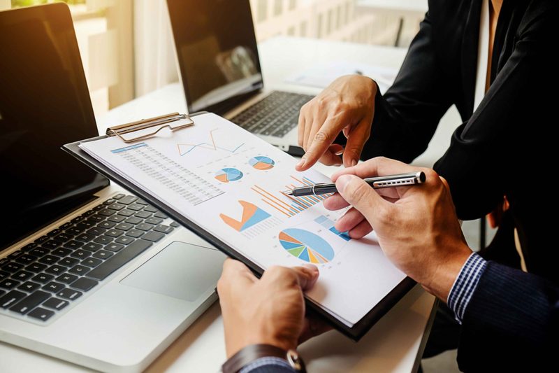 Two people in suits pointing at bar and pie charts on a clipboard